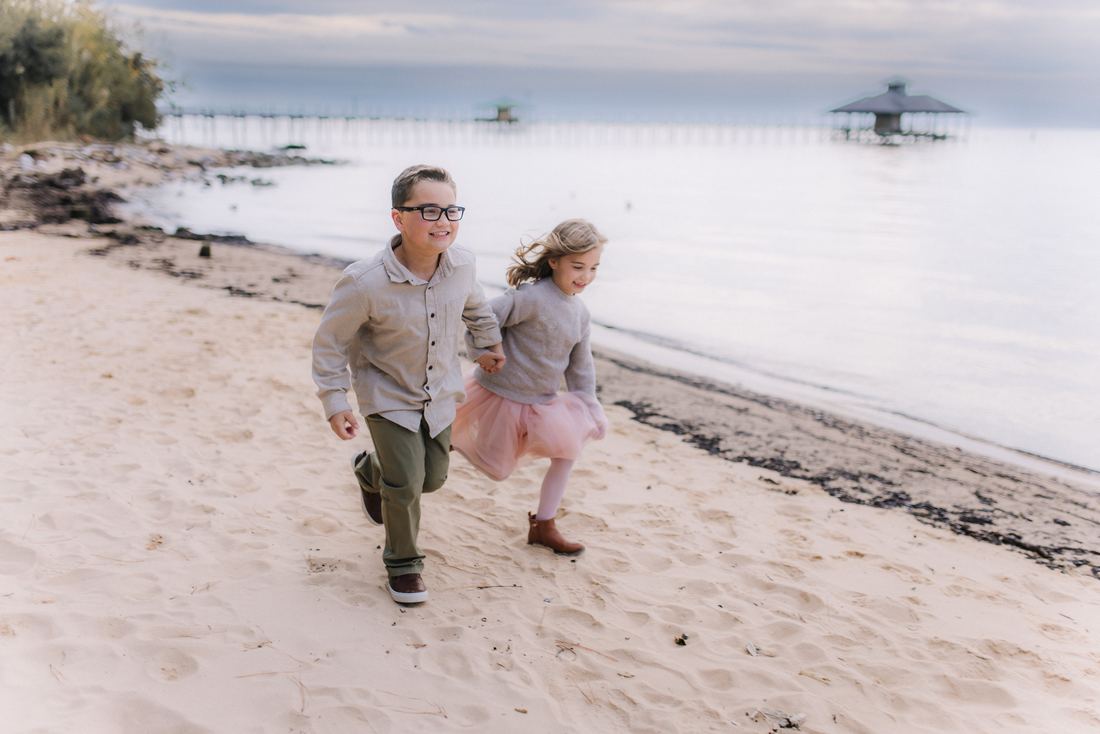 A brother and sister running hand in hand along the shoreline, the pier pavilion far out on the water behind them