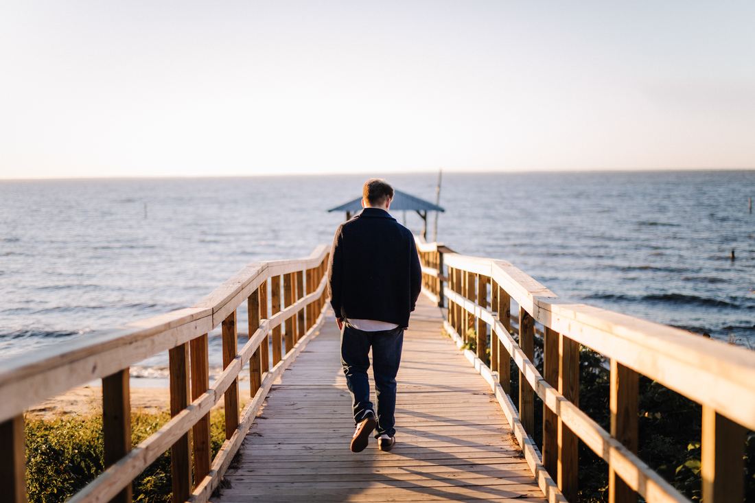A senior in a navy shawl cardigan walking away down a wooden pier toward the open bay, the water pale and wide in front of him