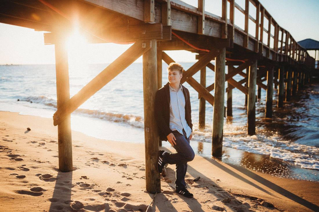 A senior leaning against a piling beneath the pier, the low sun flaring through the beams behind him