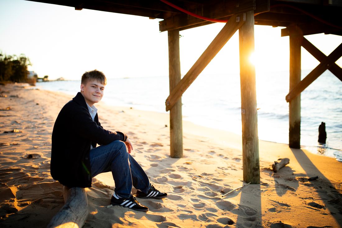 A senior seated on driftwood under the pier at sunset, looking back toward the camera as the light comes off the water