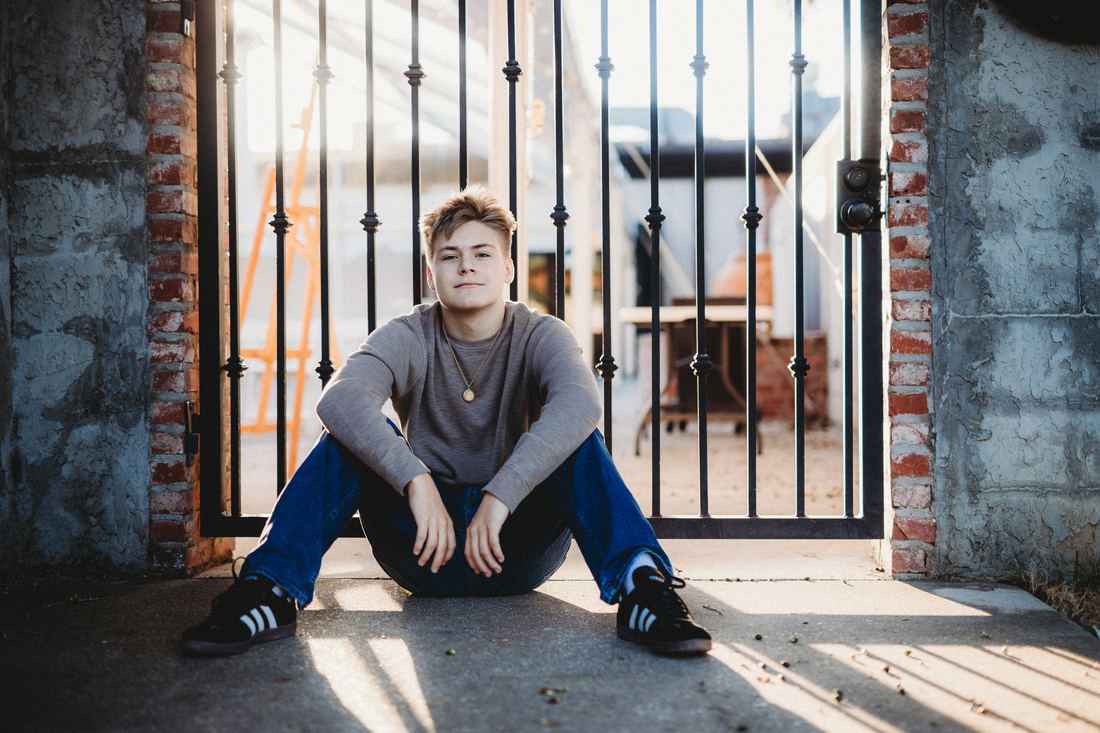 A senior in a grey thermal and jeans sitting against a wrought-iron gate set in old brick and stucco, late sun raking across the ground