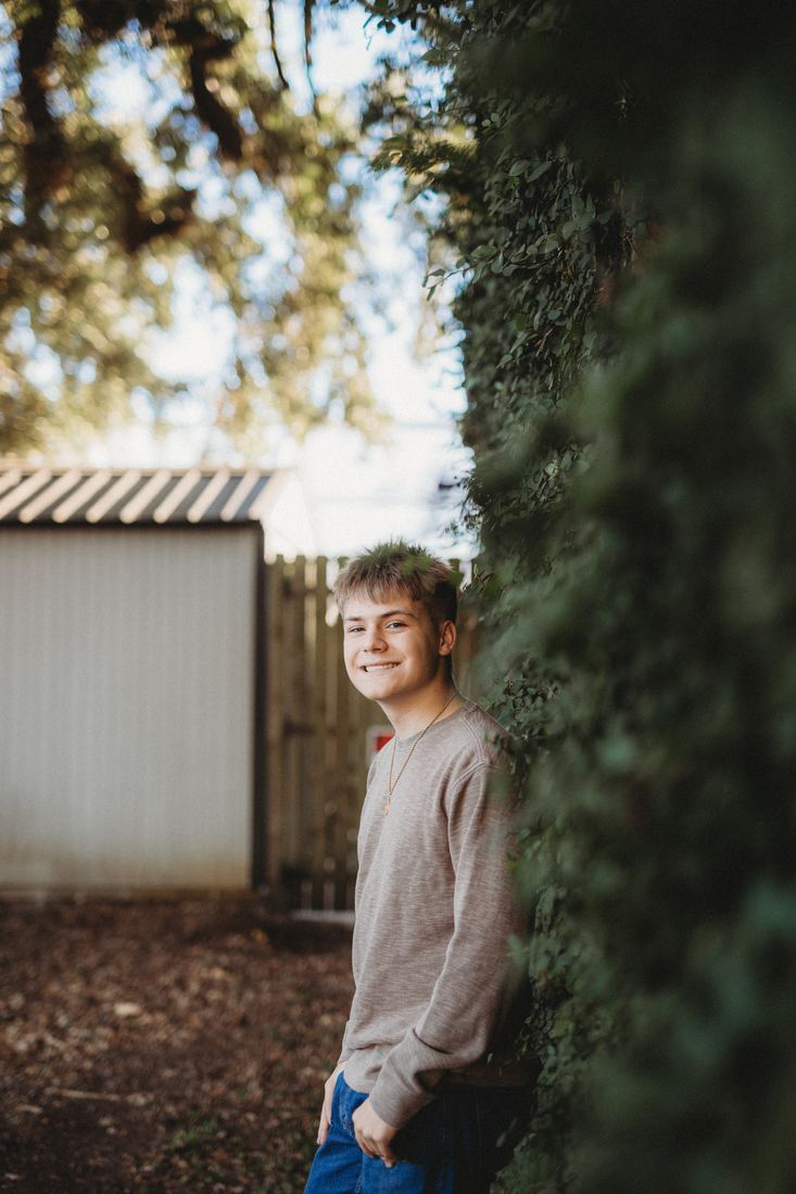 A senior standing beside a wall of ivy under a live oak, hands in his pockets, smiling toward the camera