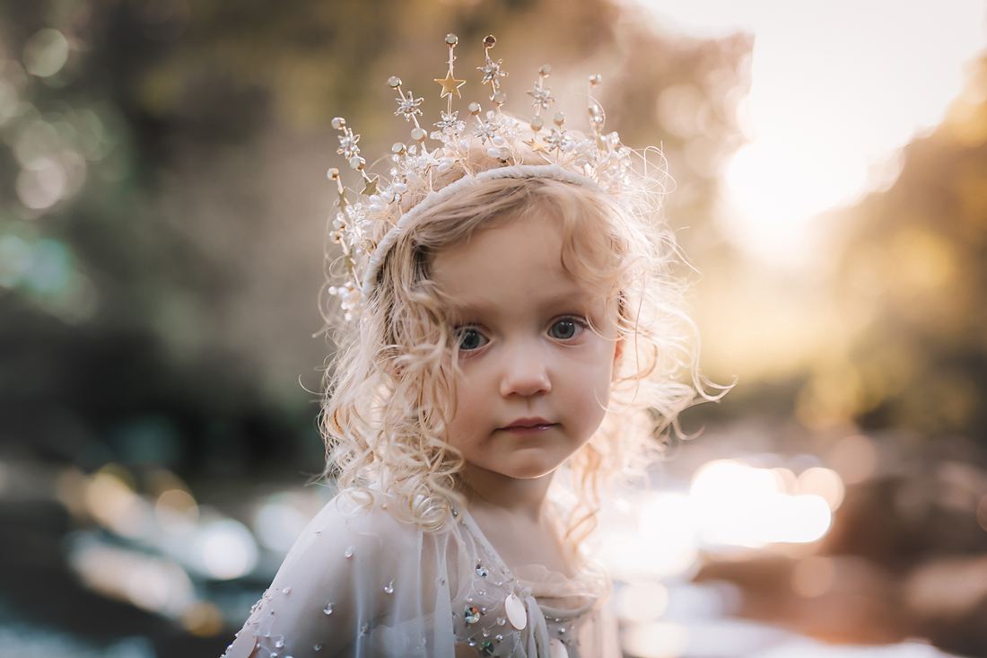 A close portrait of a curly-haired little girl in a beaded star crown and sequined tulle, the low sun glowing behind her