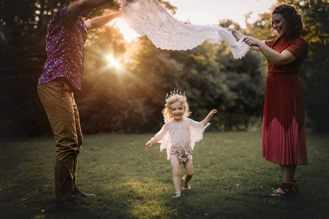 A little girl in a star crown running through the grass at sunset while her parents hold a lace shawl above her