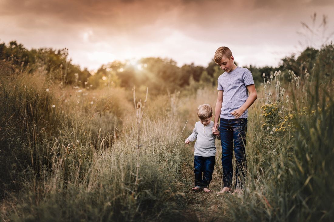 Two brothers walking hand in hand barefoot down a path through tall grass and wildflowers, the sun low behind the treeline