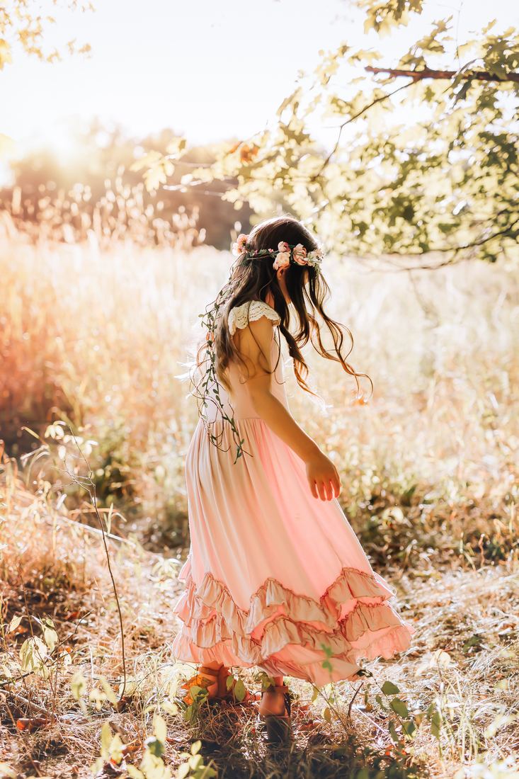 A girl in a flower crown and long blush dress turning in a sunlit field, her hair caught mid-spin