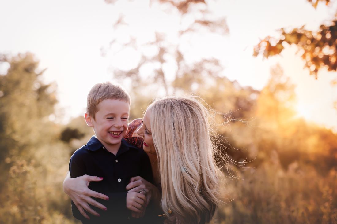 A mother leaning in to whisper to her young son, both laughing, the autumn field glowing gold behind them