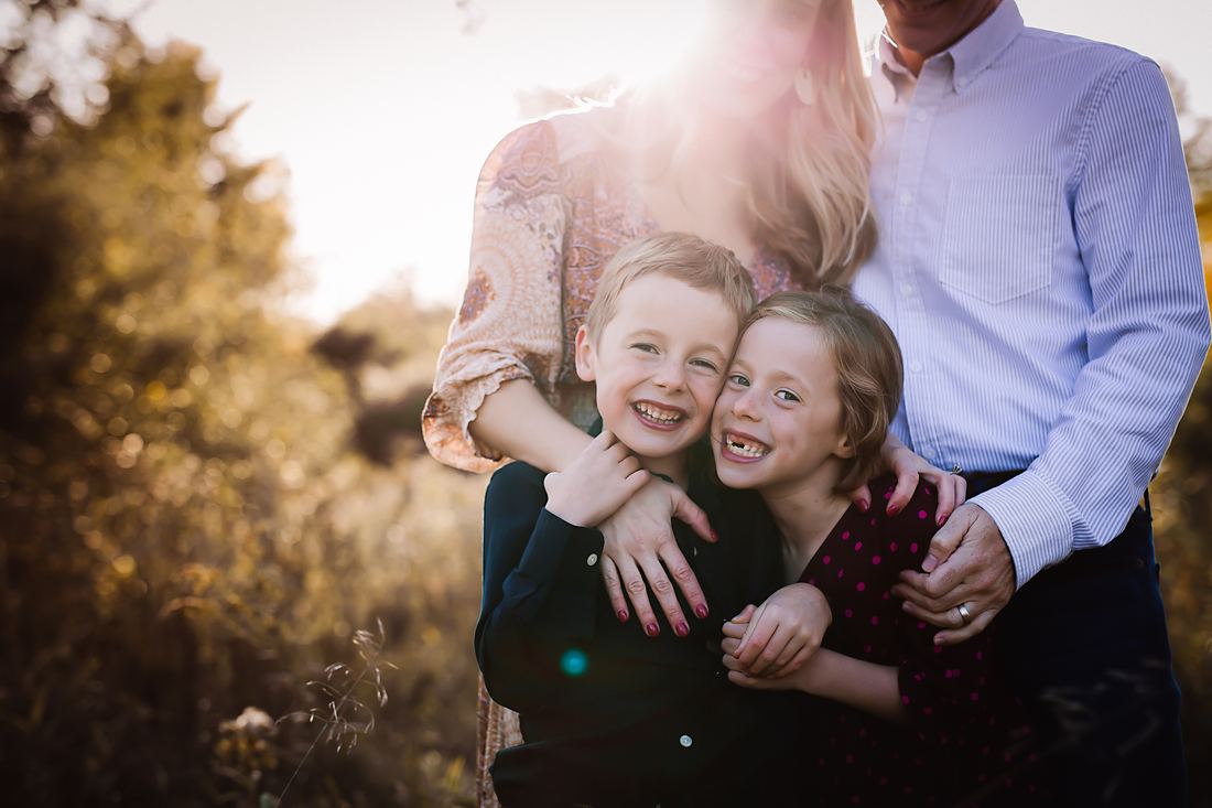 A brother and sister cheek to cheek grinning at the camera, their parents' arms wrapped around them both