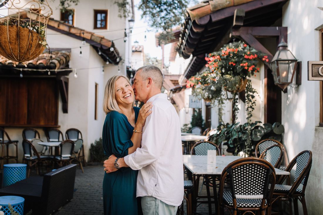 A couple holding each other and laughing in a courtyard of cafe tables, hanging baskets and gas lanterns around them