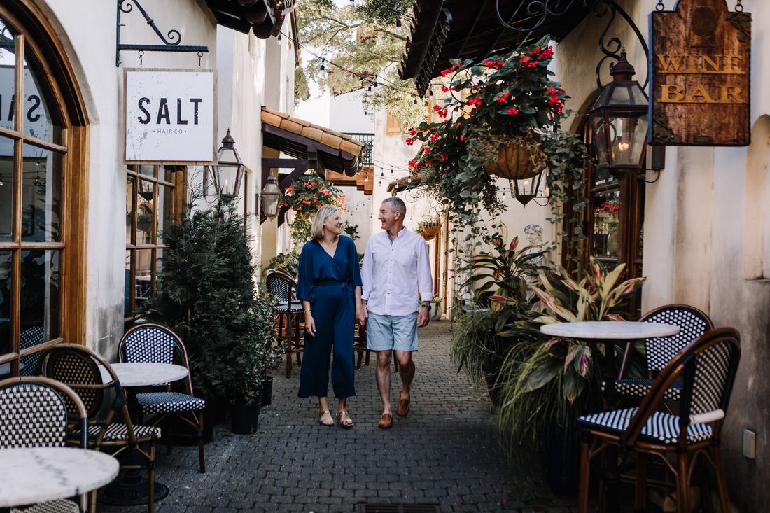 A couple walking hand in hand down a brick courtyard lined with shopfronts, string lights and red begonias overhead