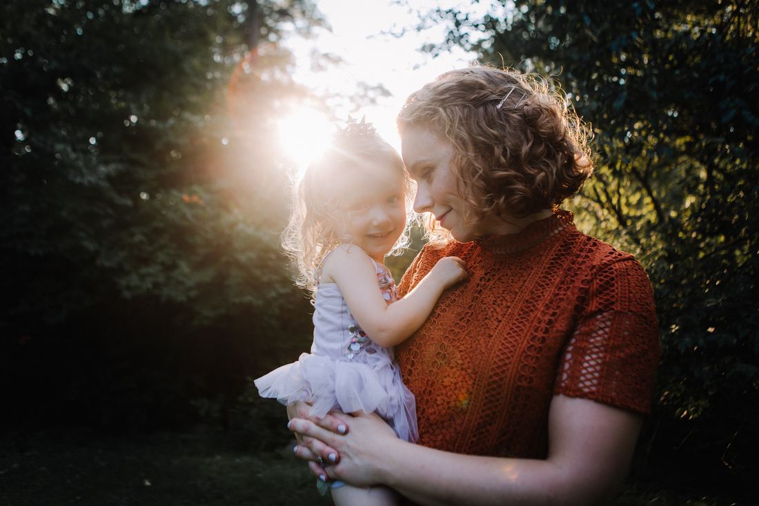 A mother holding her daughter in a star crown and tulle dress, foreheads close, the sun flaring through the trees behind them