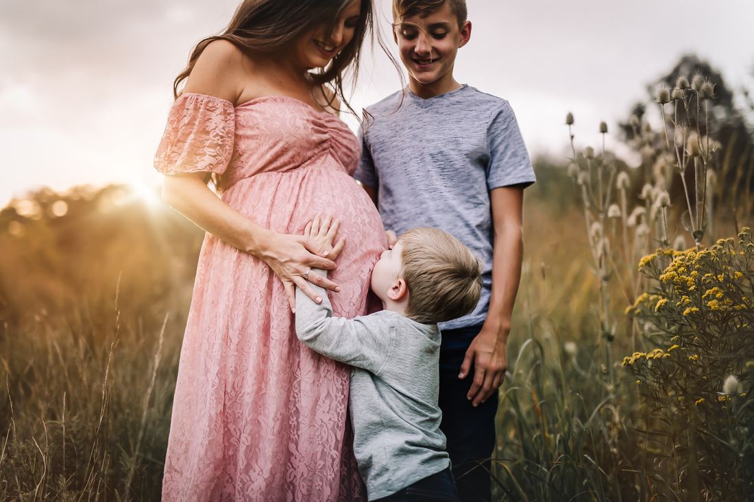 An expecting mother in a blush lace gown with her two sons, the younger one pressing his cheek against her belly
