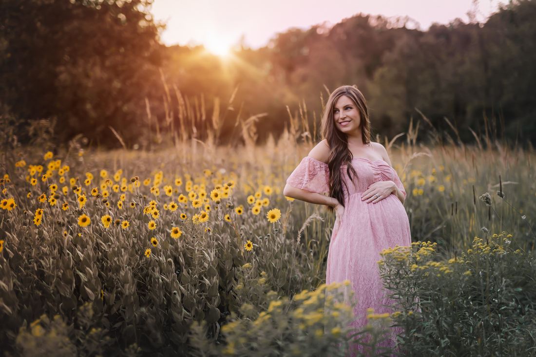 An expecting mother in a blush lace gown standing among wild sunflowers, the sun setting through the treeline behind her
