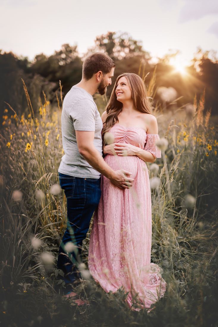 An expecting couple in tall grass at golden hour, his hands cradling her belly as she looks up at him