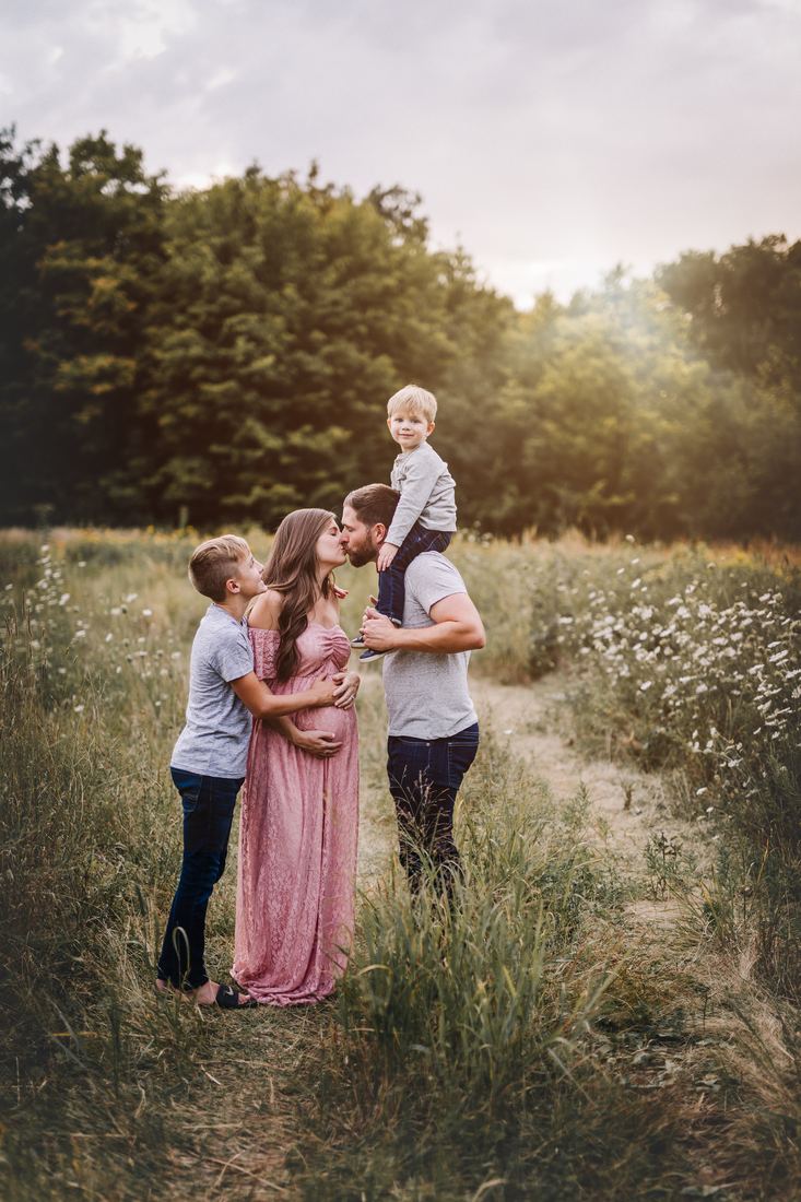 A family of four in a summer meadow, the parents kissing while their older son holds his mother's belly and the younger rides his father's shoulders