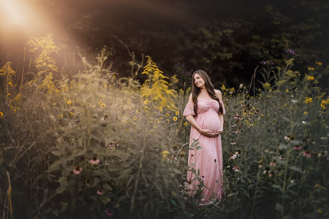 An expecting mother standing with her eyes closed in a field of goldenrod and coneflowers, sunlight breaking in from the left