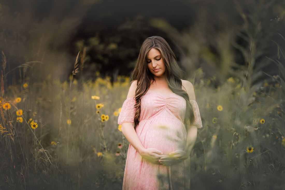 An expecting mother looking down at her belly, both hands resting beneath it, wildflowers soft all around her