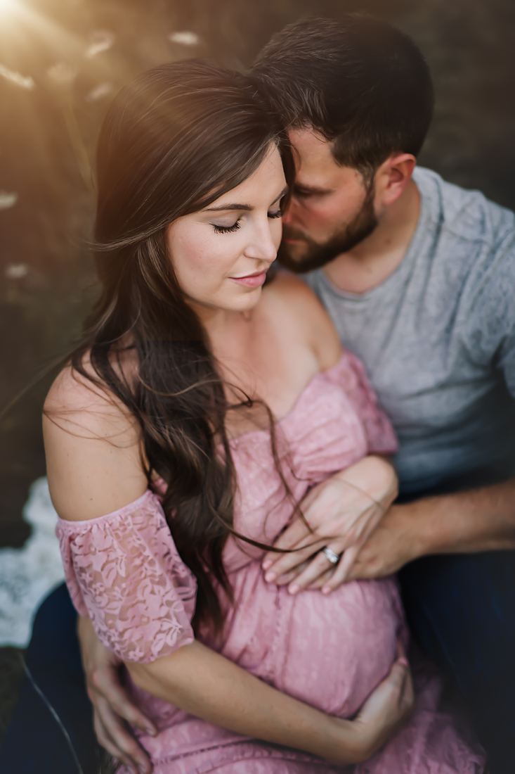 A close view of an expecting couple sitting together, his arms wrapped around her belly and his face resting against her hair