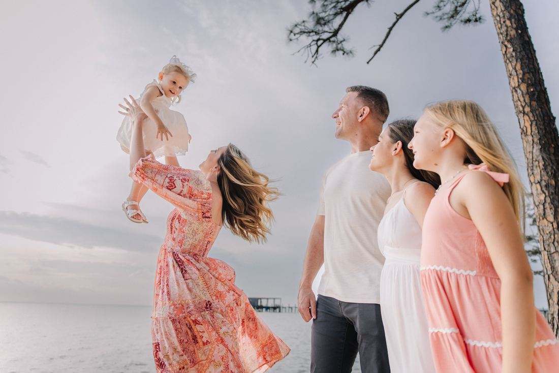 A mother lifting her toddler overhead on the shore while her husband and two older daughters watch and laugh, the bay behind them