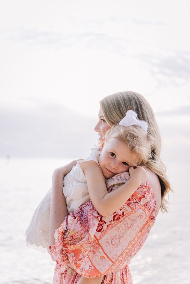 A toddler in a white eyelet dress and hair bow resting her cheek on her mother's shoulder, looking back at the camera