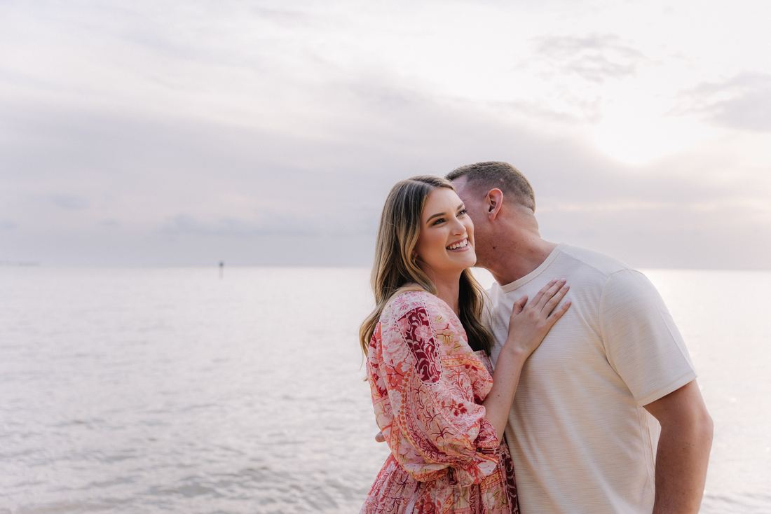 A husband kissing his wife's cheek at the water's edge while she smiles out toward the bay