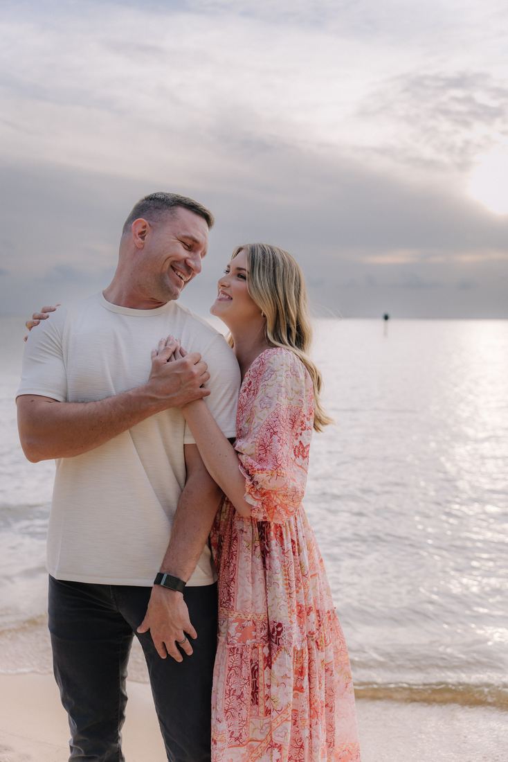 A couple laughing together on the sand, her arm around his shoulder and the evening water bright behind them