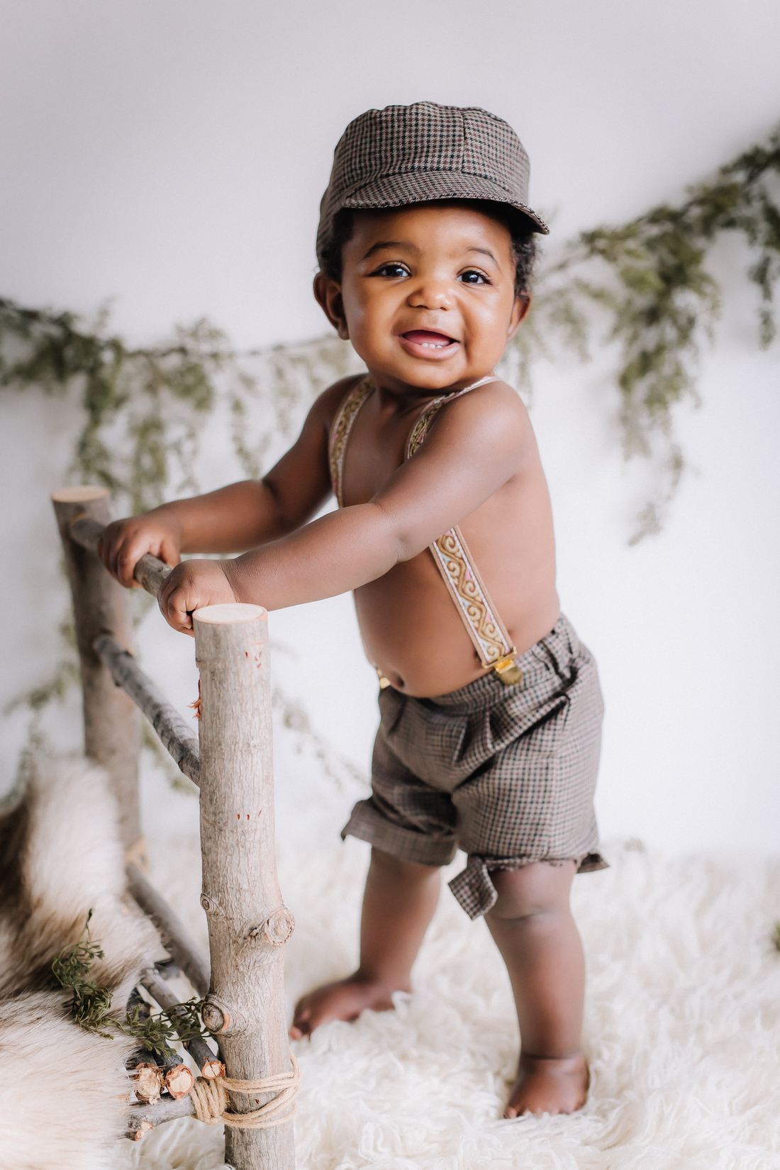 A one-year-old in a flat cap and suspenders pulling up to stand on a wooden rail