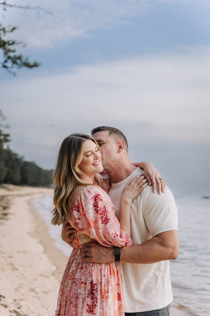 A husband holding his wife close and kissing her temple on the shoreline, pines along the sand behind them