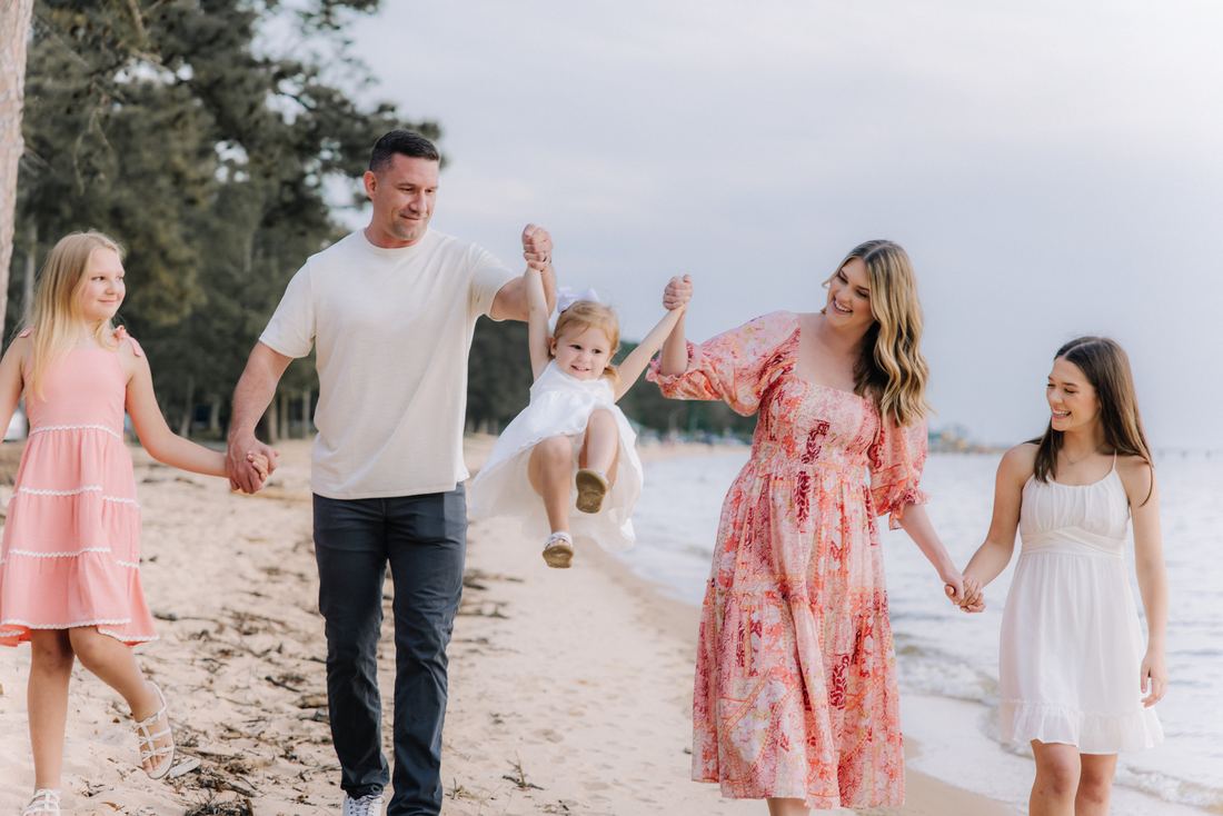 A family of five walking the shoreline, the youngest swinging between her parents' hands while her big sisters walk alongside
