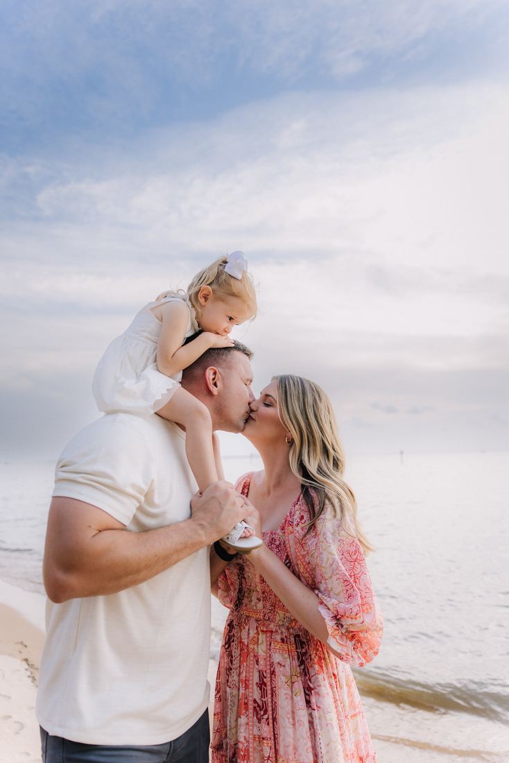 A father with his toddler on his shoulders leaning in to kiss his wife on the beach