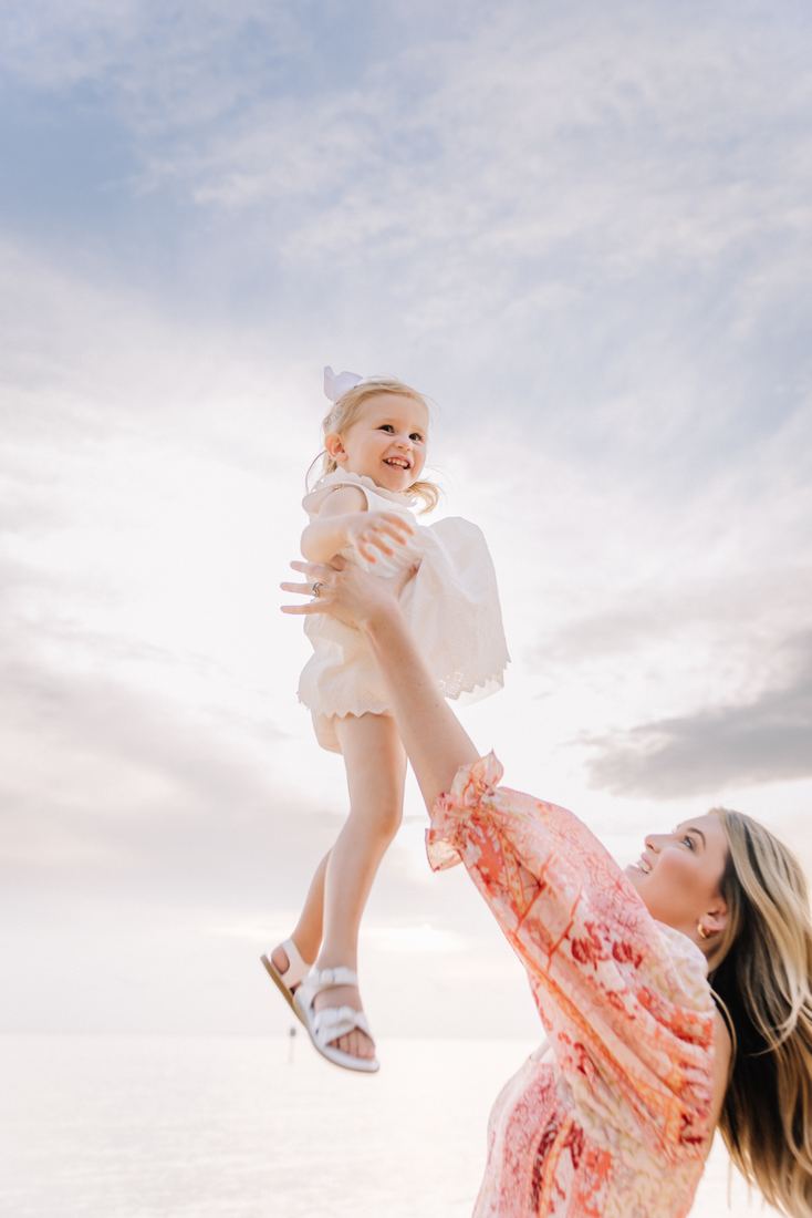 A mother swinging her laughing toddler up against a wide pastel sky