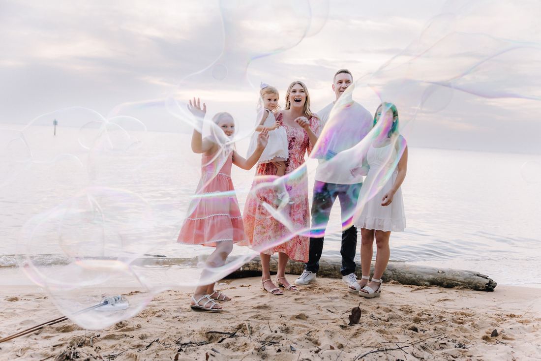 A family of five standing on driftwood at the water's edge, laughing as giant soap bubbles drift across the frame