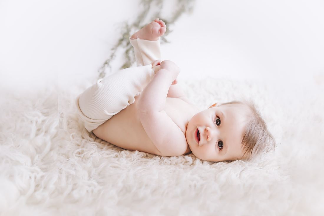 A baby on her back on a cream sheepskin, cream ribbed romper pushed up and one bare foot in the air, looking over at the camera