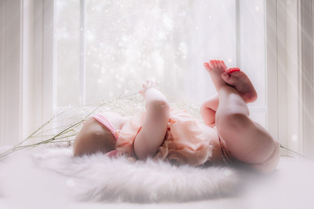 A baby in a peach ruffled romper kicking both feet up on a white fur rug, baby's breath and a bright window behind her