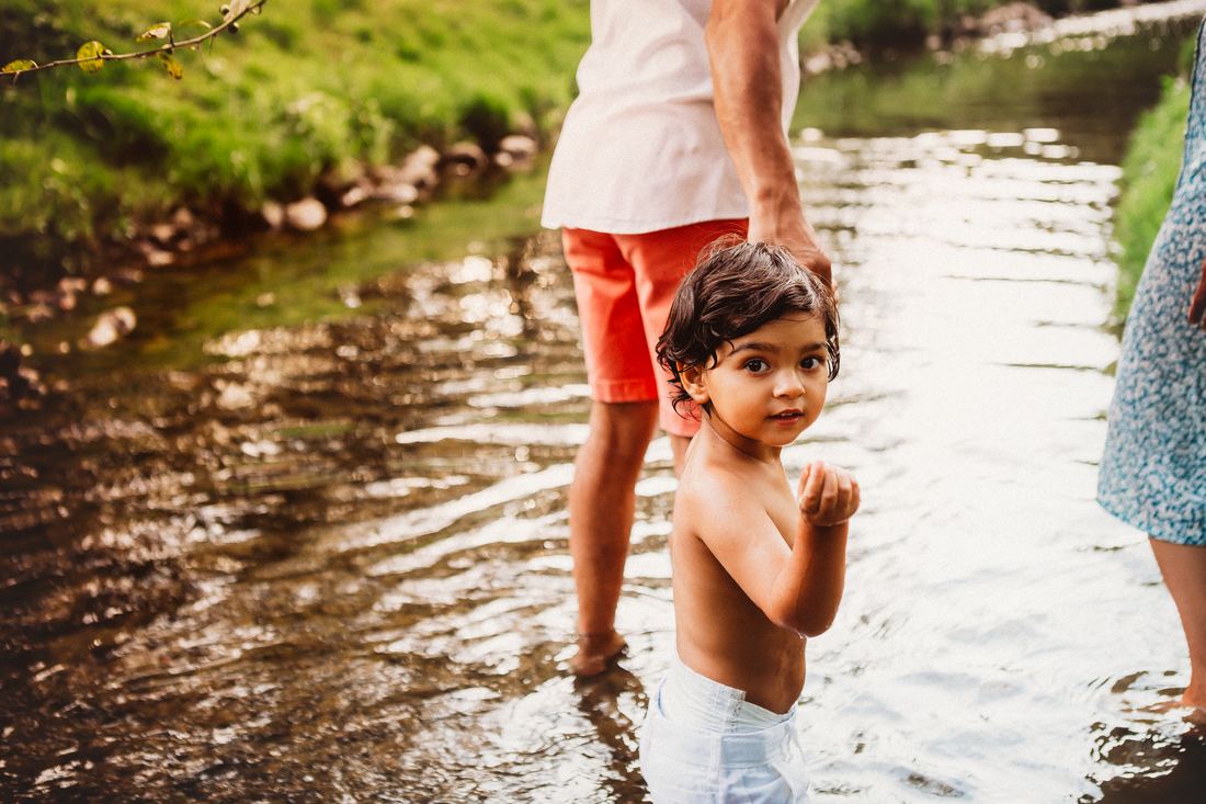 A small boy wading in a shallow creek, holding a parent's hand and looking back over his shoulder at the camera