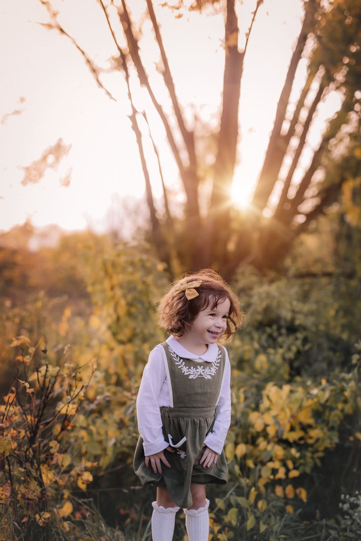 A girl in an olive pinafore and mustard hair bow laughing in an autumn thicket, the sun low through the trees behind her