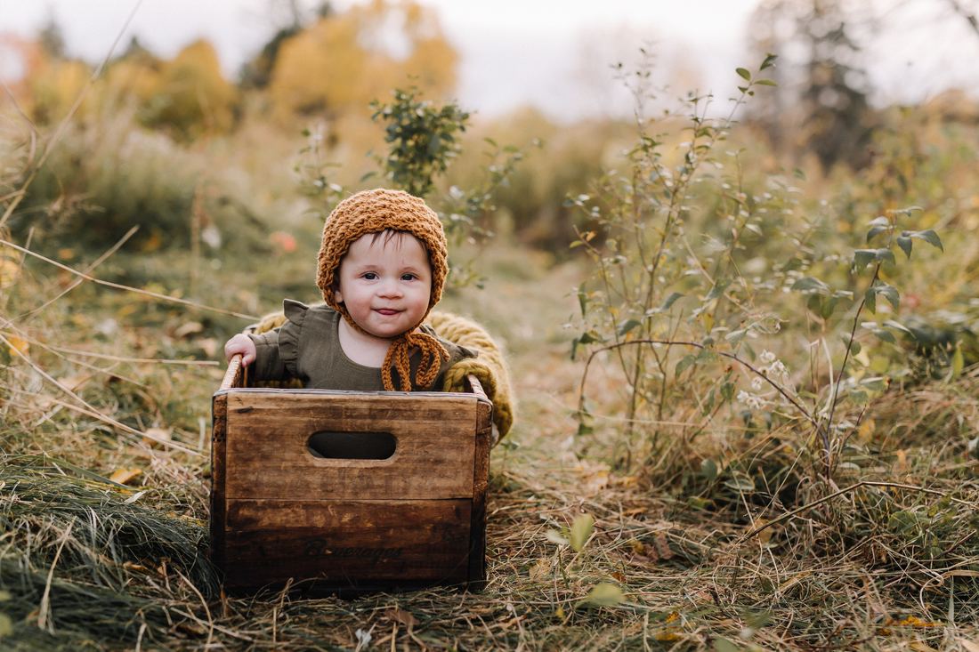 A baby in a mustard knit bonnet sitting up in a wooden crate out in a grassy autumn field