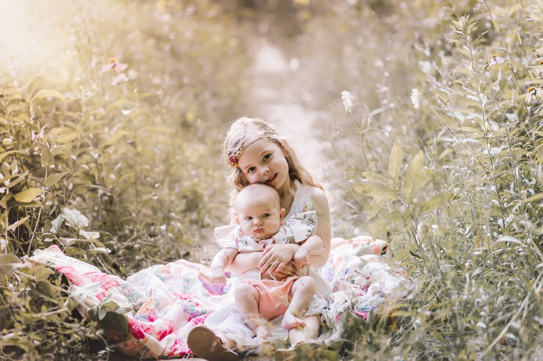 A big sister in a flower headband holding her baby sister on a patchwork quilt in tall summer grass