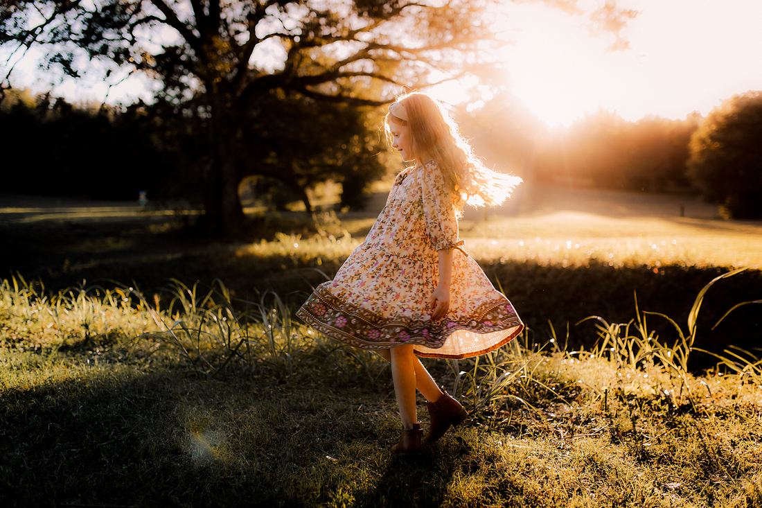 A girl in a floral dress and boots spinning under a live oak, her skirt and hair flying in the low golden light