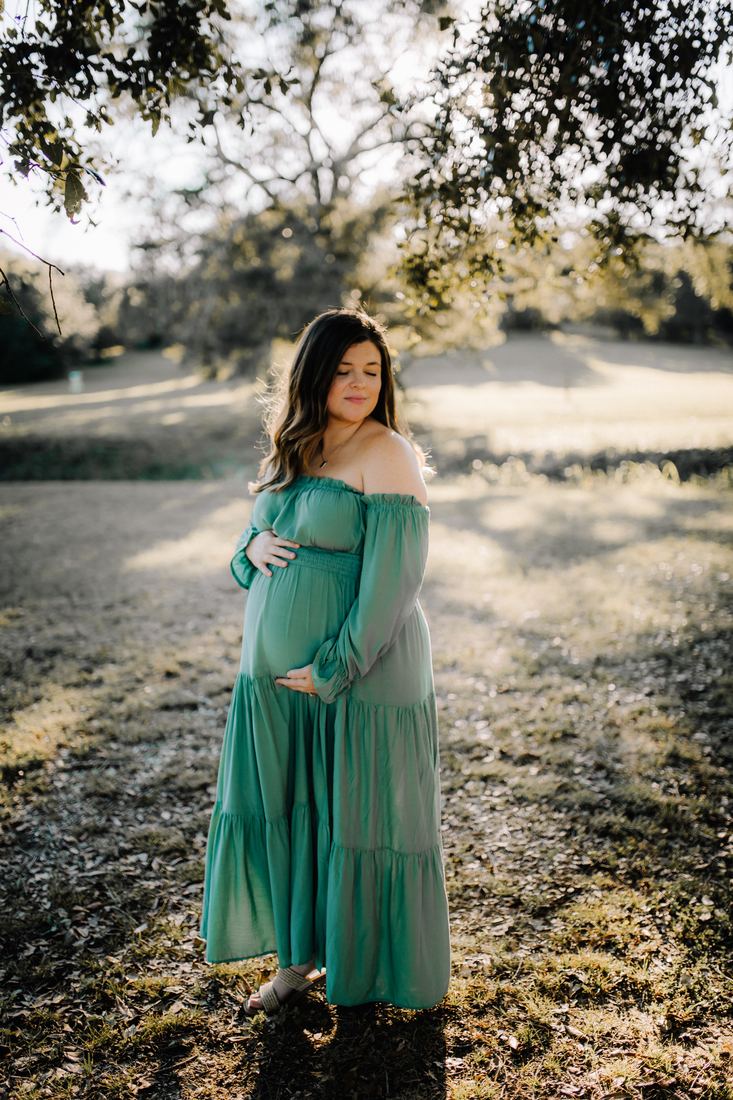An expecting mother in a green tiered dress standing under a live oak, both hands resting on her belly and her eyes closed in the dappled light