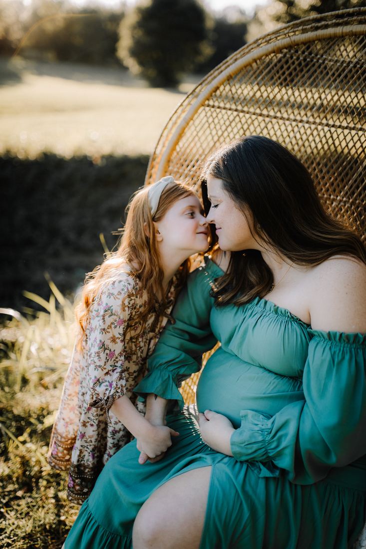 An expecting mother seated in a peacock chair in a field, nose to nose with her young daughter