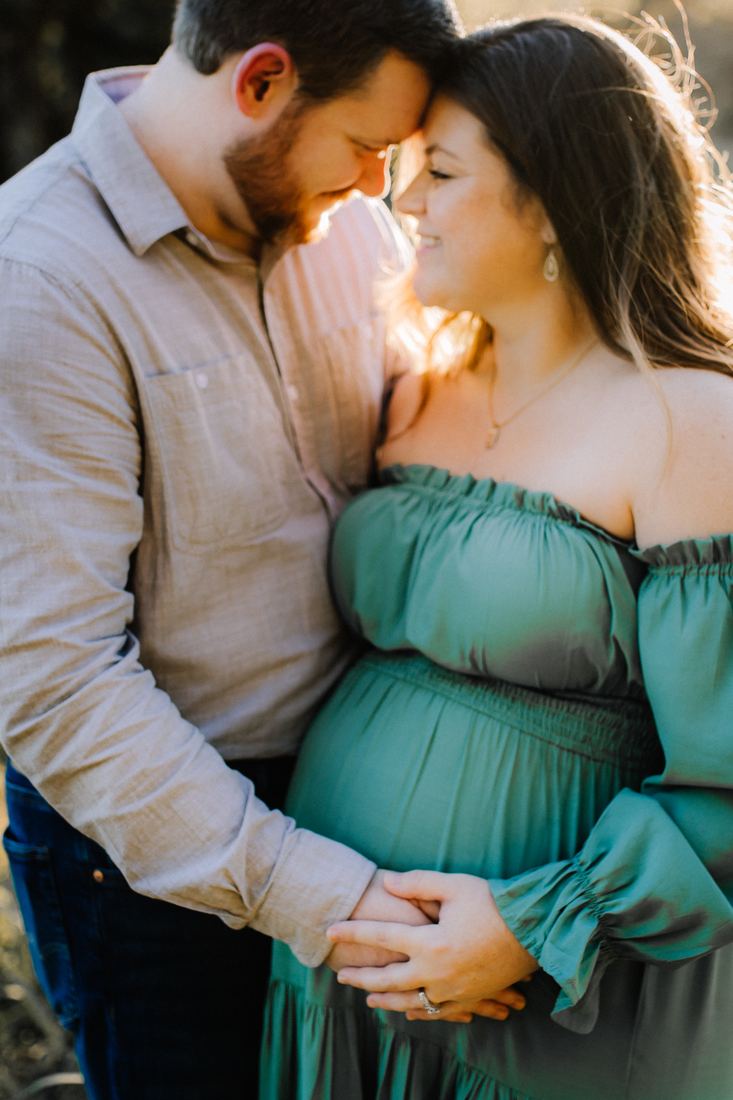 An expecting couple leaning their foreheads together, their hands joined beneath her belly in the late sun