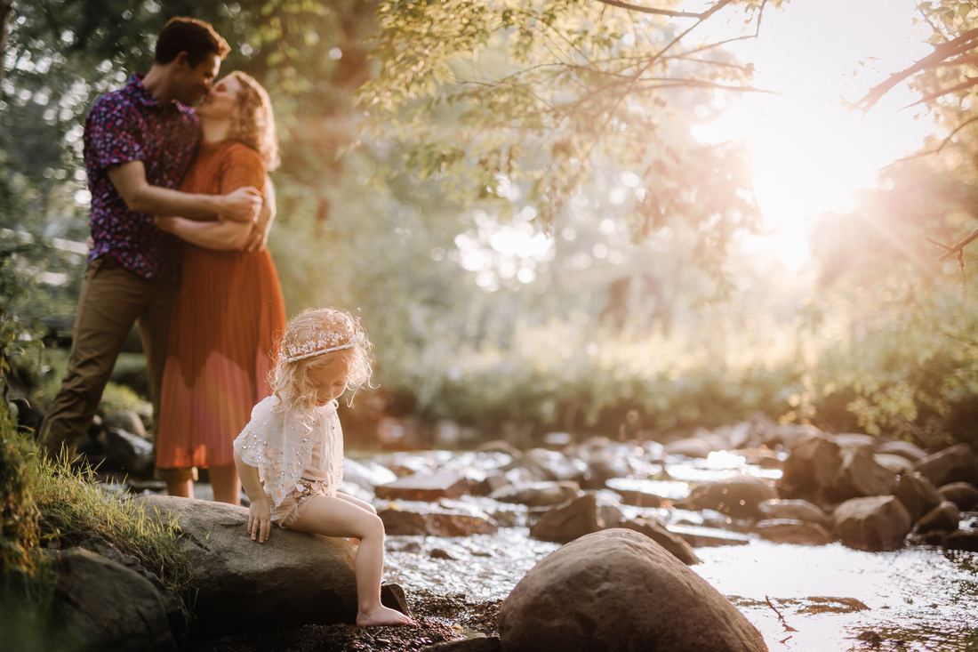 A little girl in a beaded crown sitting on a rock at the edge of a creek while her parents kiss on the bank behind her