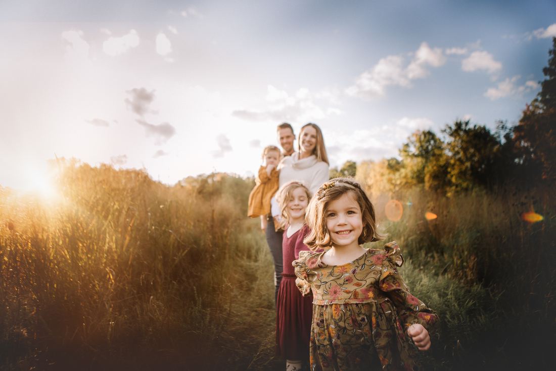 Three sisters walking a path through tall autumn grass ahead of their parents, the youngest grinning straight at the camera