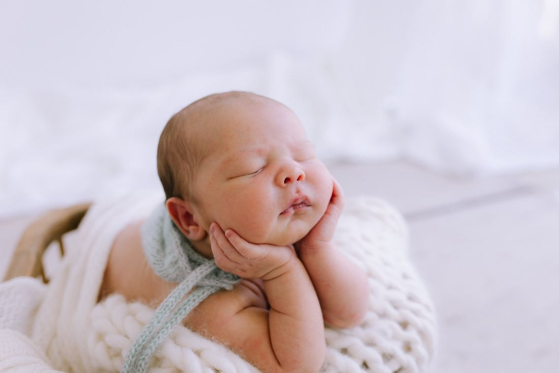 A sleeping newborn in a sage bonnet with his chin resting on both hands
