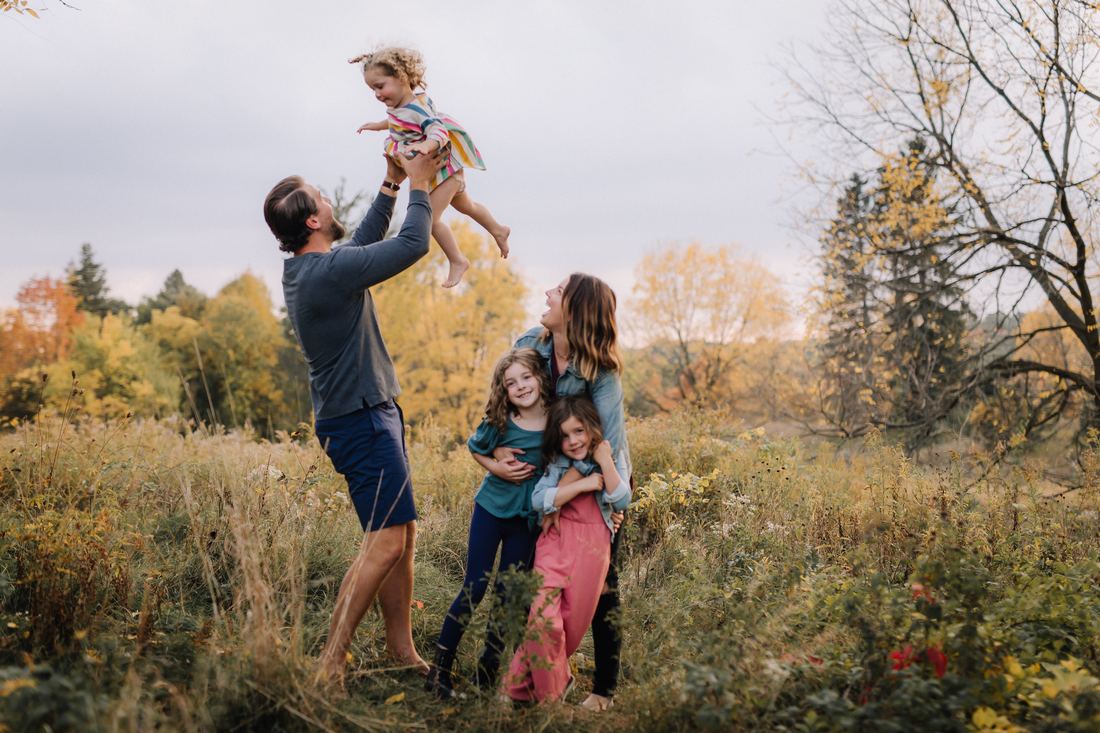 A father tossing his youngest daughter into the air in an autumn meadow while her mother holds the two older girls and laughs
