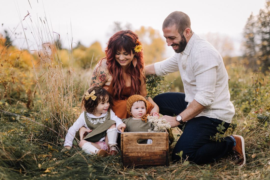Parents kneeling in the tall grass with their toddler and their baby, who sits in a wooden crate with a bundle of dried flowers