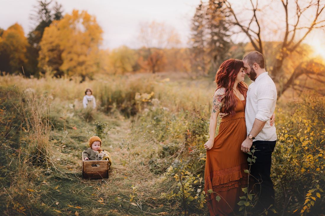 A couple standing forehead to forehead at the edge of an autumn field, their baby in a wooden crate and their older child playing further down the path