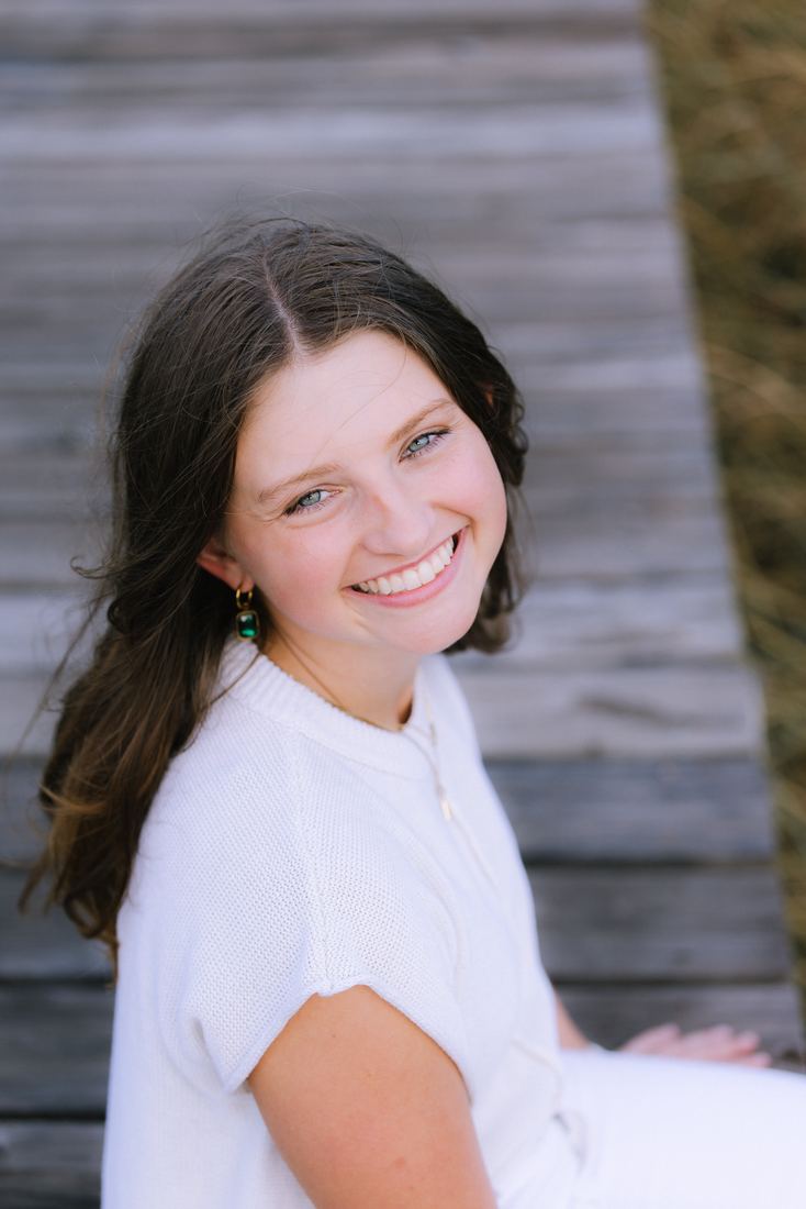 A close portrait of a senior in a cream knit top on a weathered boardwalk, turning back over her shoulder to smile at the camera