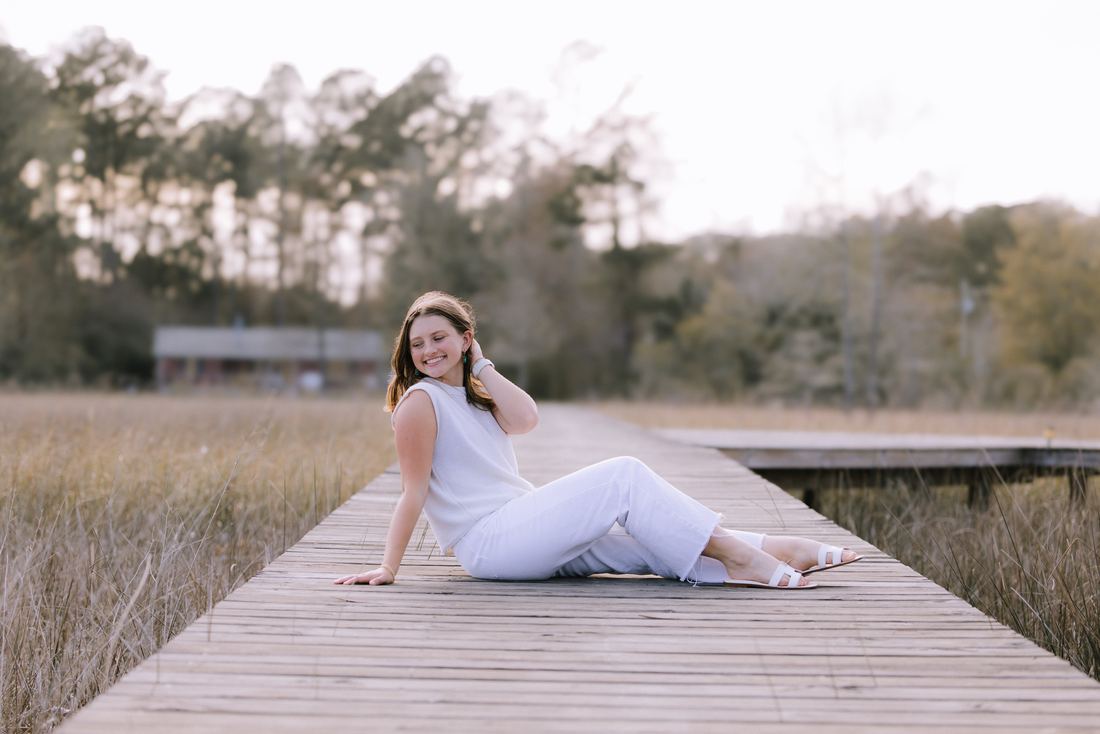 A senior sitting on a boardwalk that runs out through the marsh grass, one hand in her hair